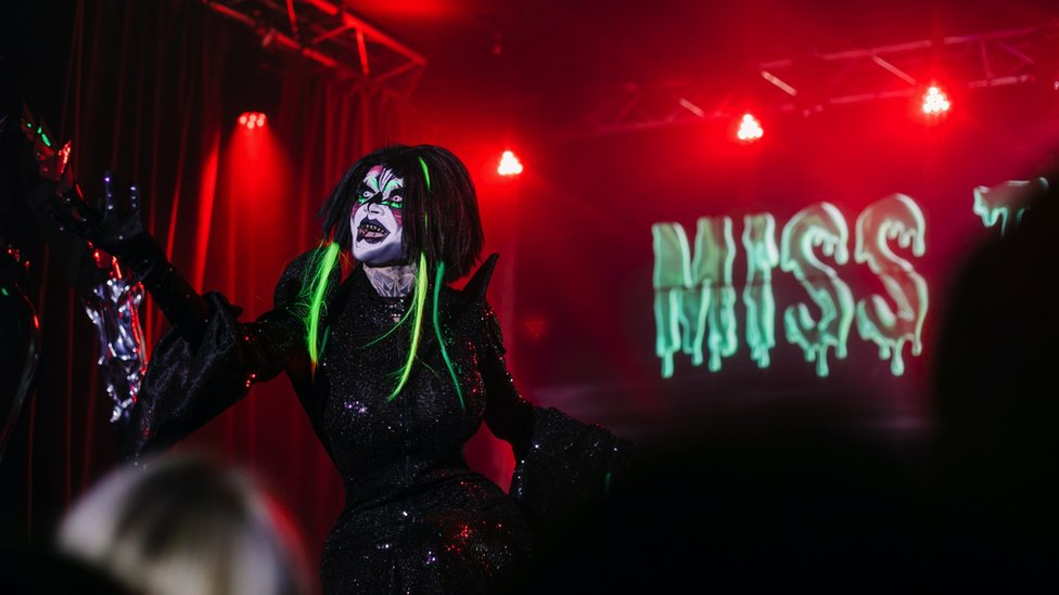 Danya on stage in a black fitted dress with white makeup and neon hair extensions