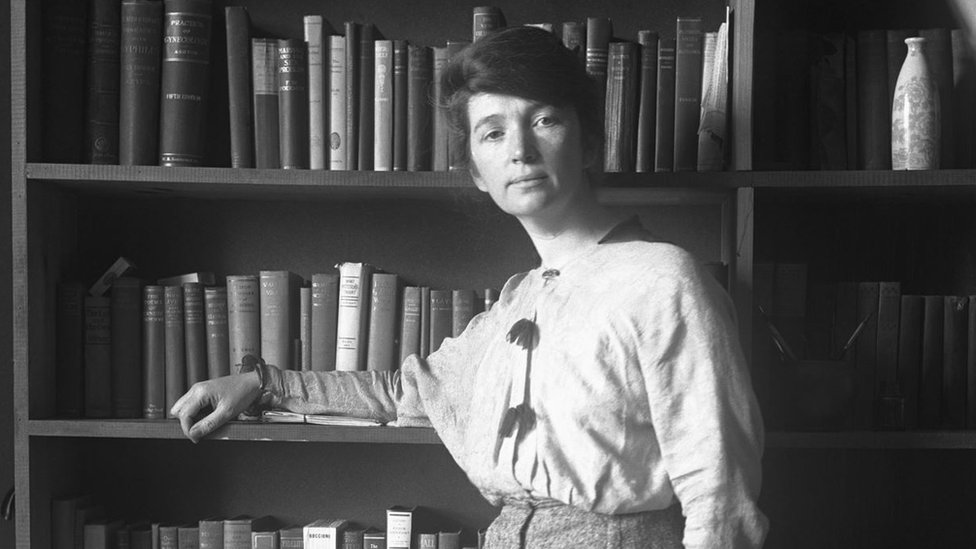 Margaret Sanger standing in front of a book shelf during her trial in Federal Court over her book The Woman Rebel