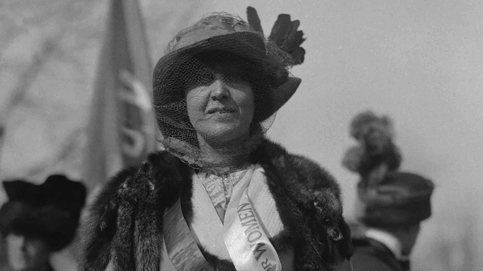 Katharine McCormick wearing a sash showing the word 'Women' with a banner being held in the background