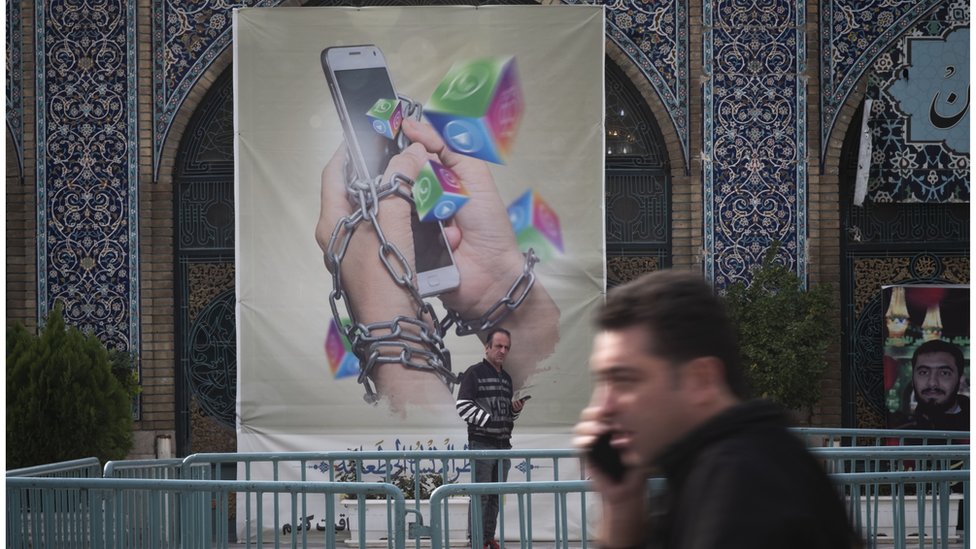 An Iranian man holding his smartphone looks on while standing under an anti-social networking banner in Tehran Grand Bazaar on 3 December 2022