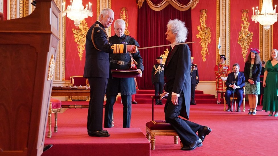 King Charles lays a sword on the shoulder of Brian May while knighting him at Buckingham Palace
