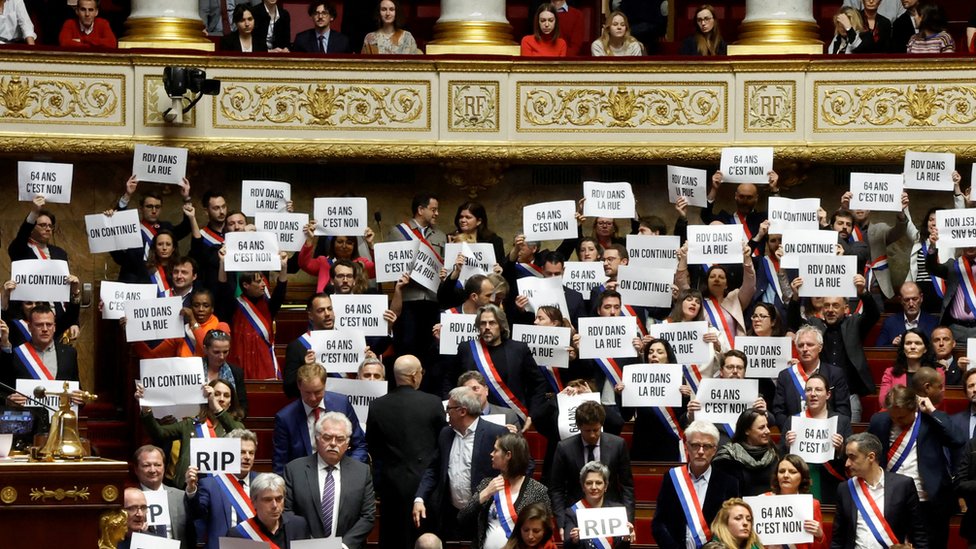 Supporters of the no-confidence motions hold placards in the National Assembly reading 'retrait', '64 ans c'est non' and 'on continue'