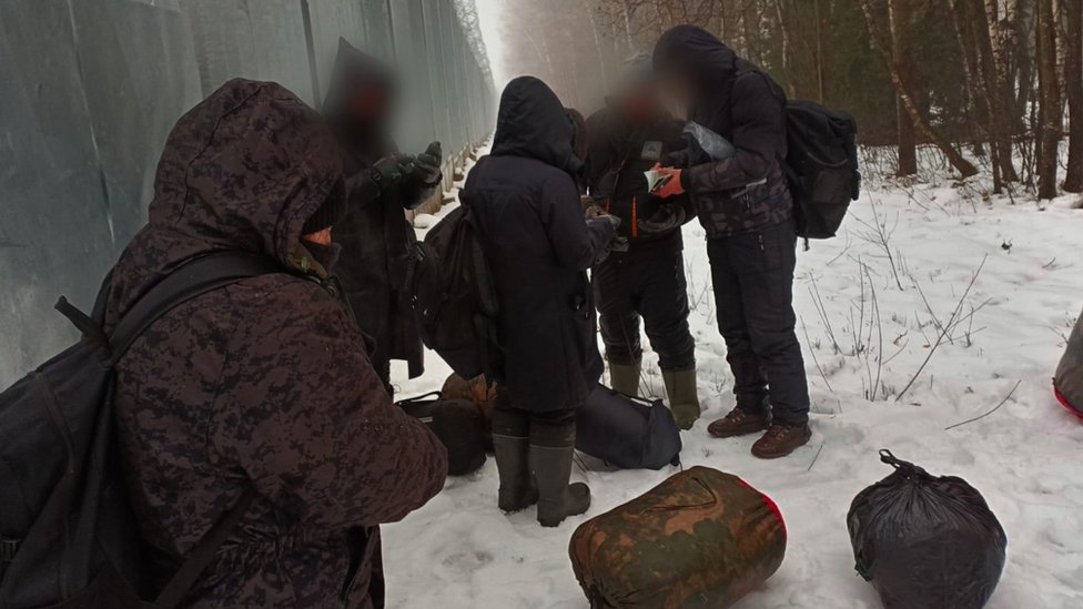 Migranti i Evropska unija: Smrtonosno putovanje - „Ugasili su šest cigareta na mom telu“ 2 A group of migrants in coats and boots look at their phones in snowy conditions