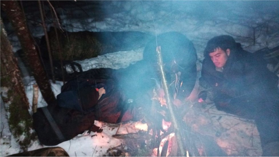 Four people huddle around a camp fire, two are sleeping while another two try to light a fire