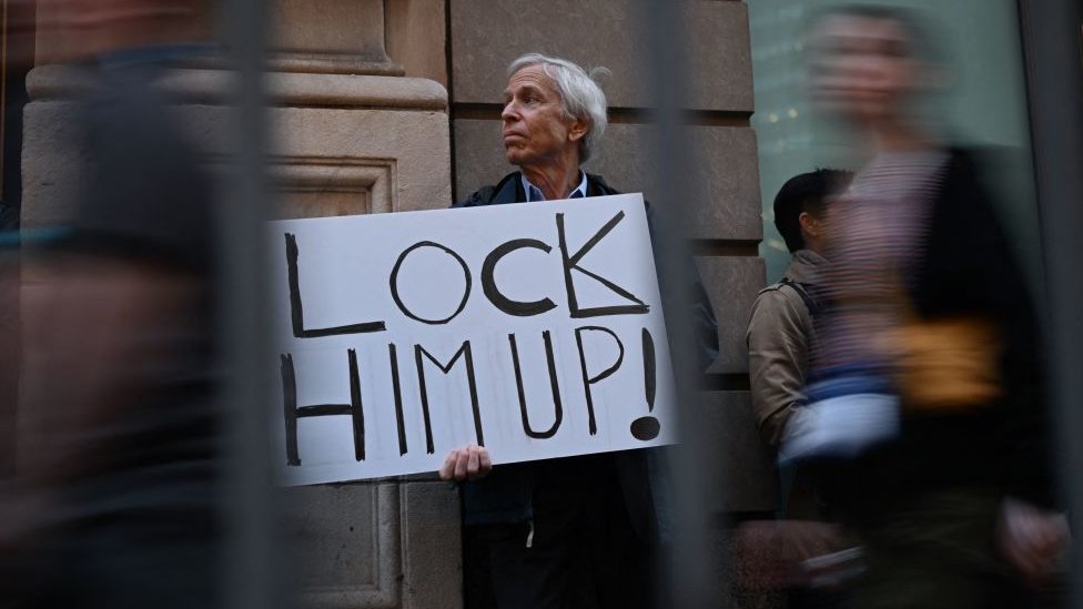 Amerika i Donald Tramp: Tramp se vratio na Floridu i poručio - „Ova zemlja odlazi dođavola" 6 A protester holds a sign reading "Lock Him Up!" as he stands outside of a media area near Trump Tower