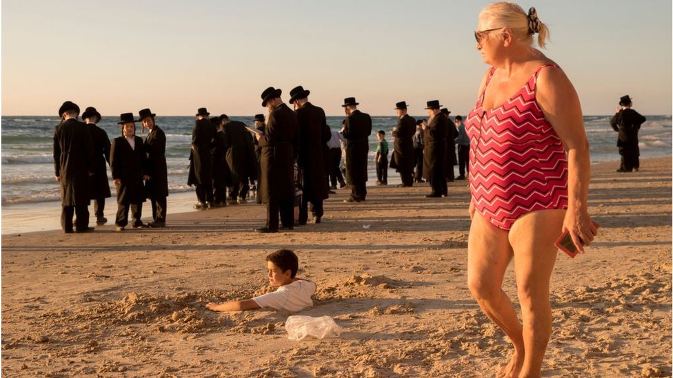 Izrael i Palestinci: Šta je sledeće za „paralelne svetove“ u zemlji? 1 A woman in a bathing suit watches a group of Orthodox Jews on a beach near Tel Aviv