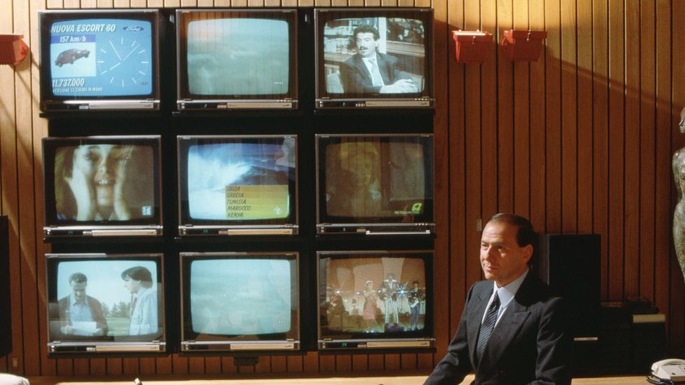 A younger Silvio Berlusconi sits in an ornate wood-panelled office with nine very large (for 1986) televisions arrayed in a grid on the wall behind him