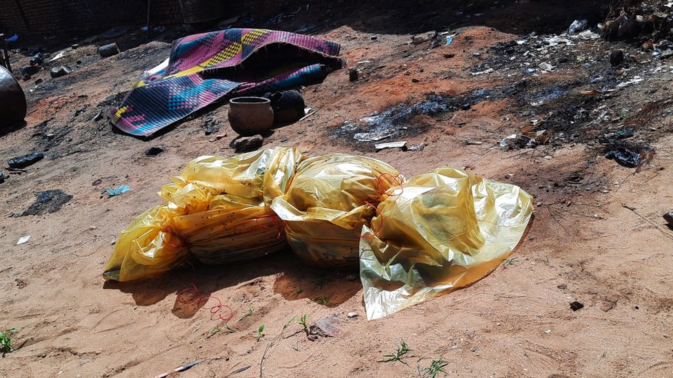 Yellow plastic bag wraps a dead body and covers it in the middle of sand and rubble. Colourful tapestry and some pots made of clay are seen scattered on floor.
