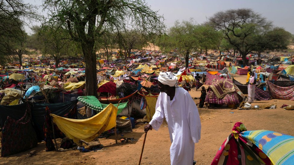 Sudanese man holds cane and walks amid thousands of colourful tents in middle of desert.