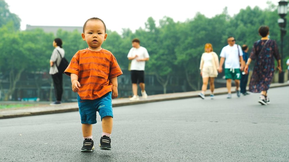 A young boy in an orange T-shirt looks into the camera