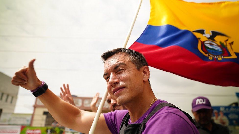 Ecuadorian presidential candidate Daniel Noboa at a campaign event. He is giving a thumbs up to the crowd and holding a large Ecuadorian flag which billows over his left shoulder. He is also wearing a bullet-proof vest.
