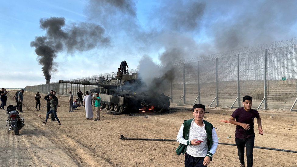 Izrael i Palestina: Desetine Izraelaca taoci Hamasa, stotine mrtvih na obe strane u napadima, „u ratu smo", kaže Netanjahu 1 Palestinians react as an Israeli tank burns on the Israeli side of Israel-Gaza perimeter fence (7 October 2023)