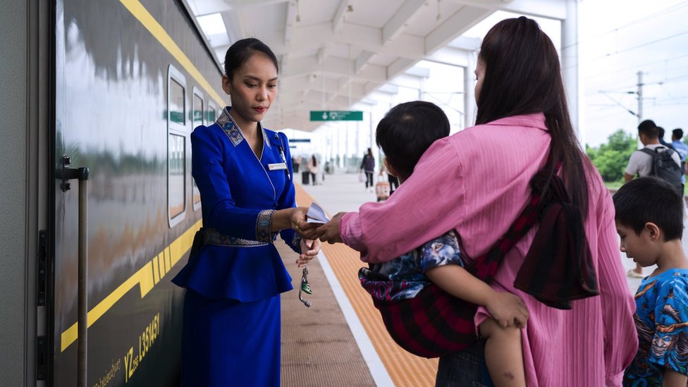 Kina: Kako je inicijativa Pojas i put vredna više biliona dolara promenila svet 7 A woman and two young boys with her waiting to board the train at the Vientiane high speed railway station, the woman handing her ticket to a female inspector in uniform