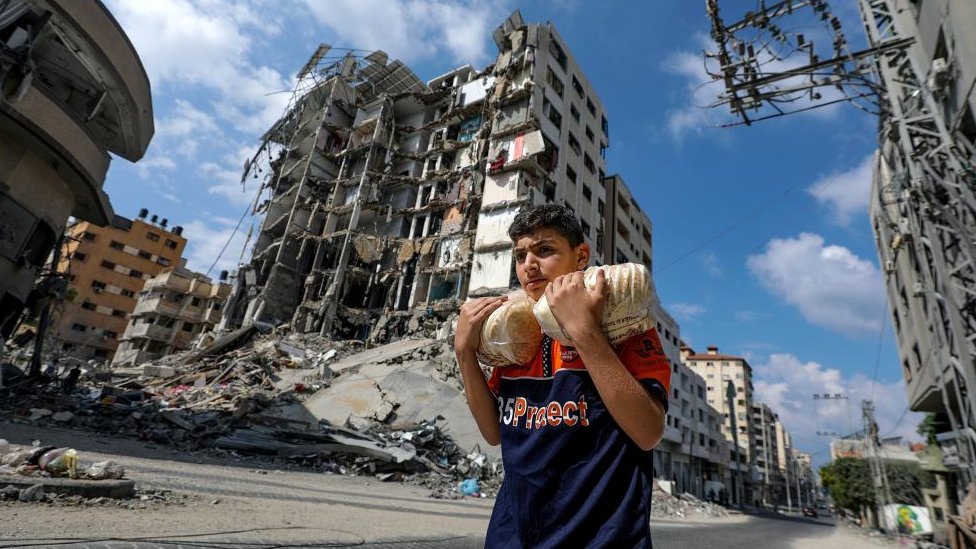 Izrael i Palestinci: Hoće li se druge zemlje umešati u sukob, BBC novinari odgovaraju na vaša pitanja 1 A Palestinian youth carrying bread walks past buildings destroyed in recent Israeli strikes in Al Remal, in Gaza City, 16 October 2023