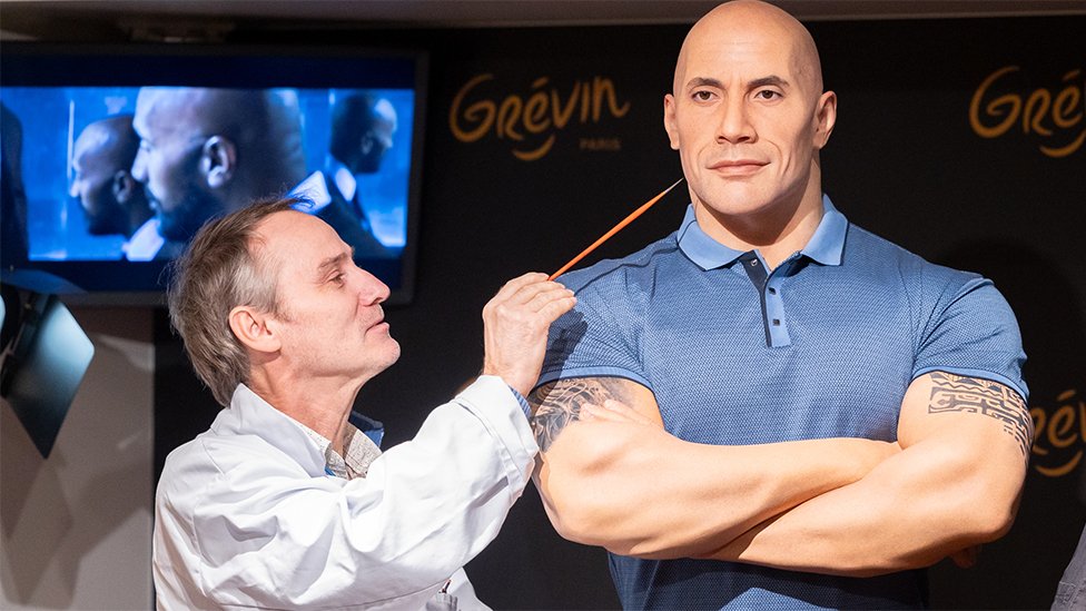 The Dwayne Johnson wax figure at Musee Grevin in France, with the painter putting a paint brush on the model. The painter is wearing a white jacket, looking towards the wax work of The Rock. The model is showing Dwayne Johnson as bald, and wearing a light blue polo shirt. The figure's arms are crossed, showing off its biceps, which are painted with swirling tribal patterns in black ink. The model looks lifelike but the skin tone is quite light.