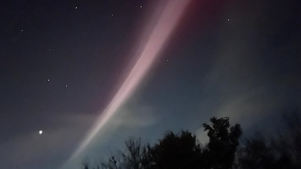Thin stream of purple and white light in the night sky with some white cloud and bright stars.