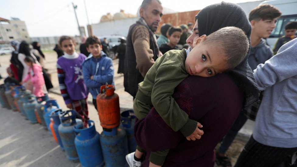 Izrael i Palestinci: Hamas optužuje Izrael za kršenje primirja, puštanje talaca „odloženo" 1 A woman carries a child as Palestinians gather to fill liquid gas cylinders, during a temporary truce between Hamas and Israel, in Rafah in the southern Gaza Strip