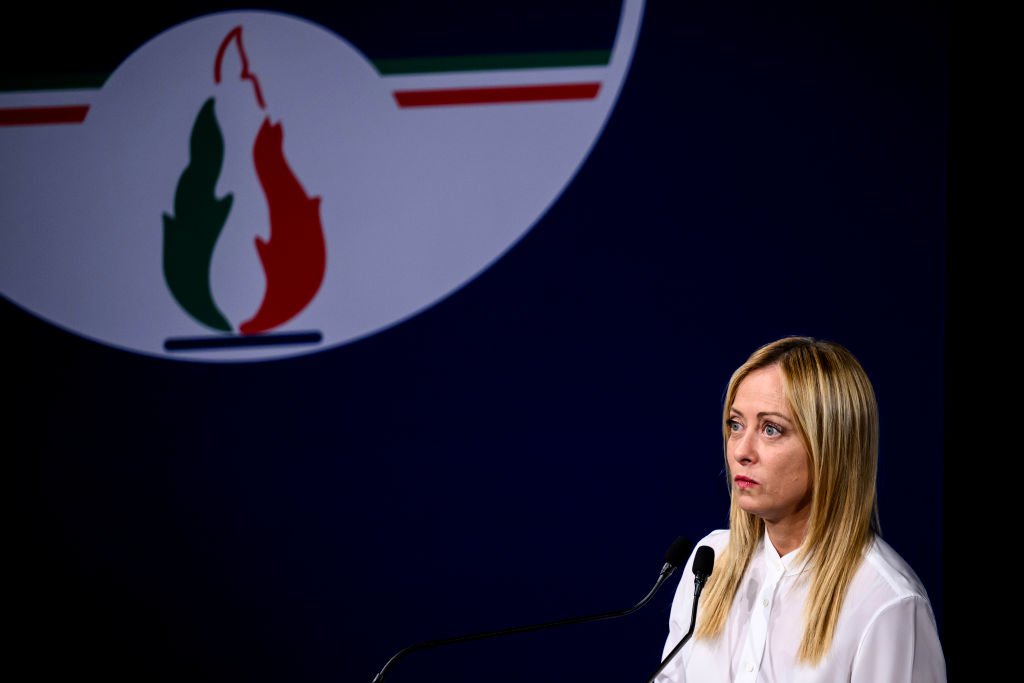 Giorgia Meloni standing next to a large sign of her party's logo, the tricolour flame