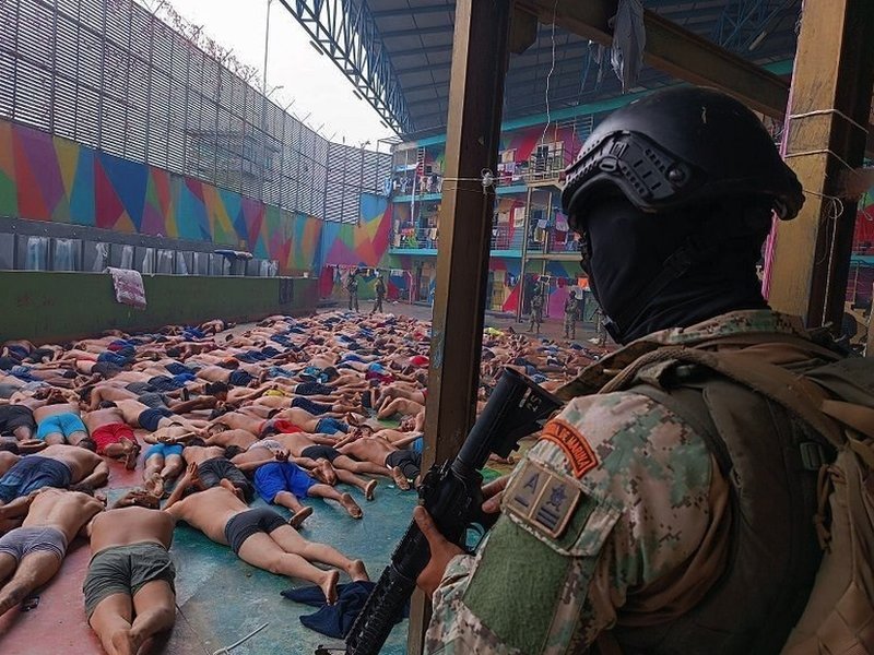 A soldier stands guard over dozens of prisoners, lying face down in a prison courtyard