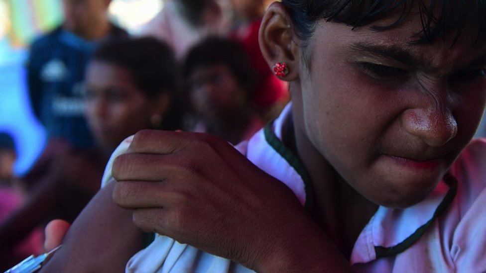 A Rohingya refugee girl scrunches her eyes as a health worker injects an MMR vaccine dose into her arm