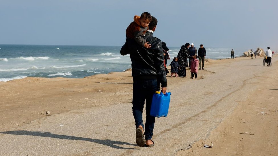 Izrael i Palestinci: Hamas izneo kontraponudu za početak mirovnih pregovora - prvo žene i stari, pa povlačenje izraelske vojske 1 A man carries a child while holding a container, as displaced Palestinians, who fled their houses due to Israeli strikes, take shelter in a tent camp amid the ongoing conflict between Israel and Hamas, near the border with Egypt in Rafah in the southern Gaza Strip