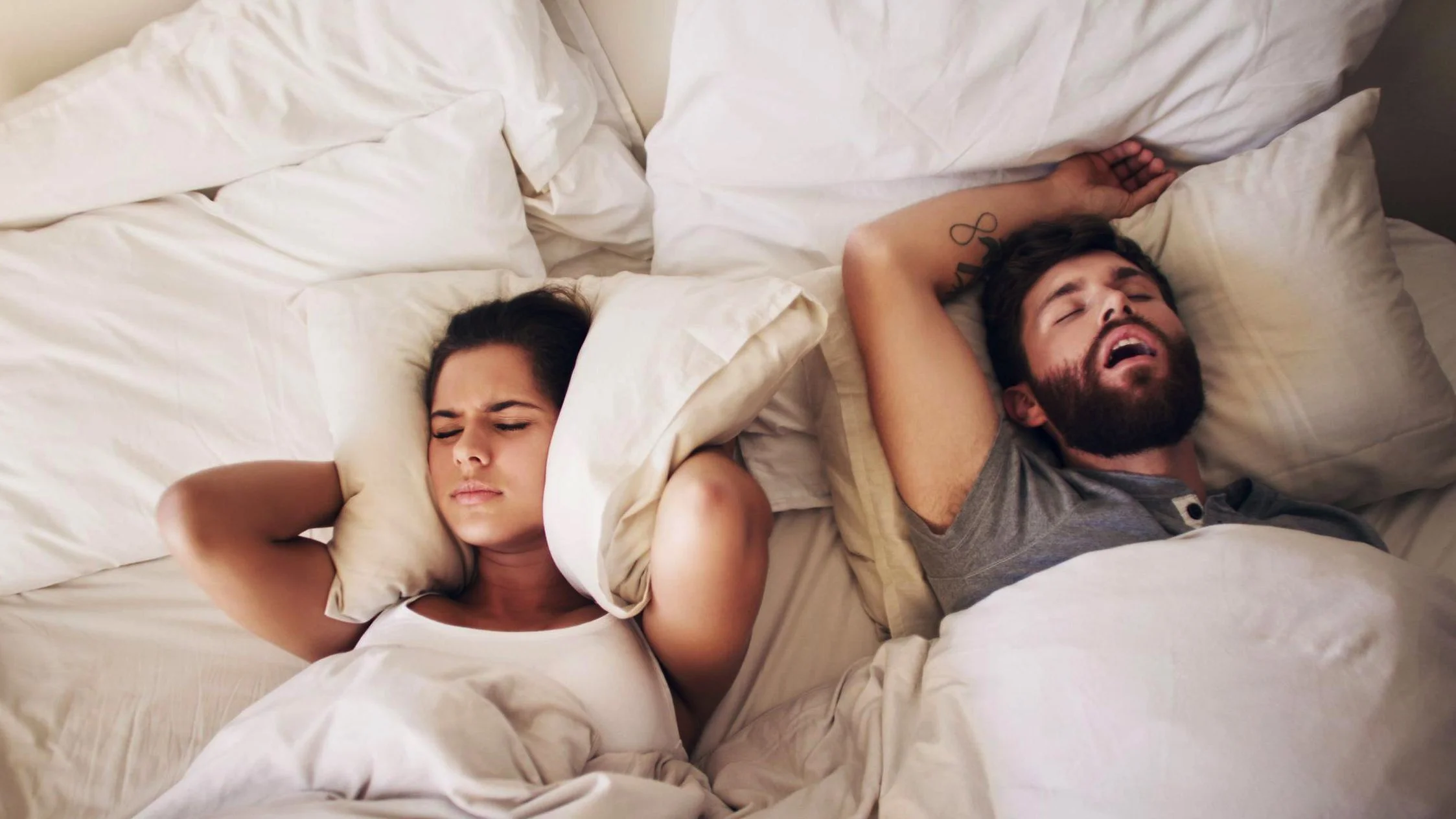 A woman covers her ears with a pillow while a man snores while sleeping