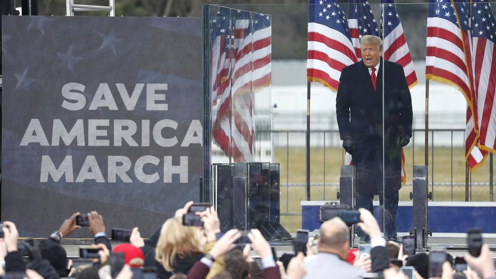 Trump standing at microphone next to sign reading Save America March