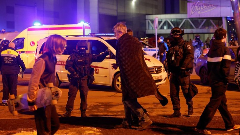 Men cover themselves with blankets while walking past Russian law enforcement officers, who stand guard near the burning Crocus City Hall concert venue