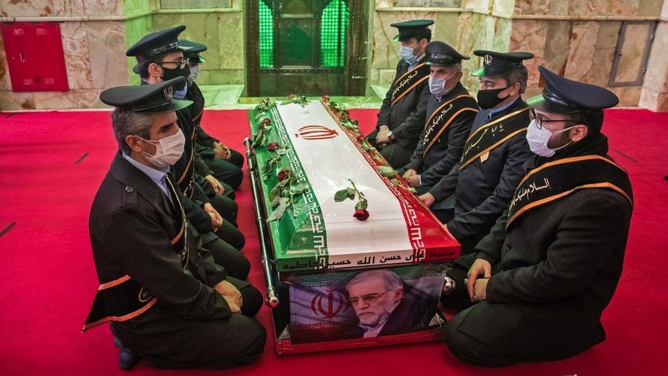 Men in uniform kneel by the coffin of the dead scientist, which has his image and an Iranian flag draped over it