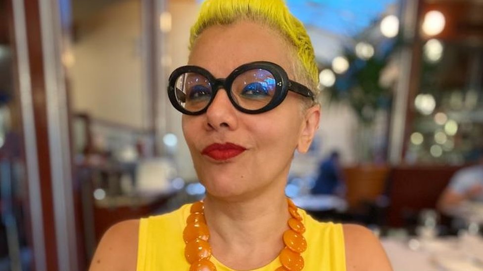 Mona, with bright yellow cropped hair and wearing glasses with thick, dark rims