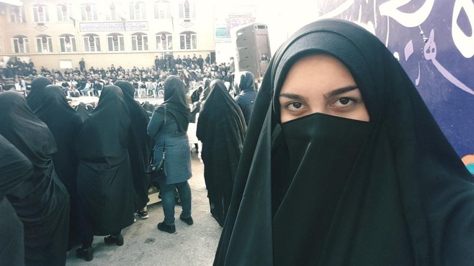 Ribell wearing a hijab that shows her eyes and forehead, standing in front of a lot of other people wearing a hijab