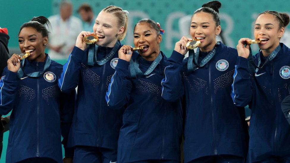 Olimpijske igre u Parizu 2024: Šta se dešava iza kulisa 2 Američki ženski gimnastičarski tim, s leva na desno: Simon Bajls, Džordan Čils, Džejd Keri, Sunisa Li i Hili Rivera drže zlatnu medalju u zubima