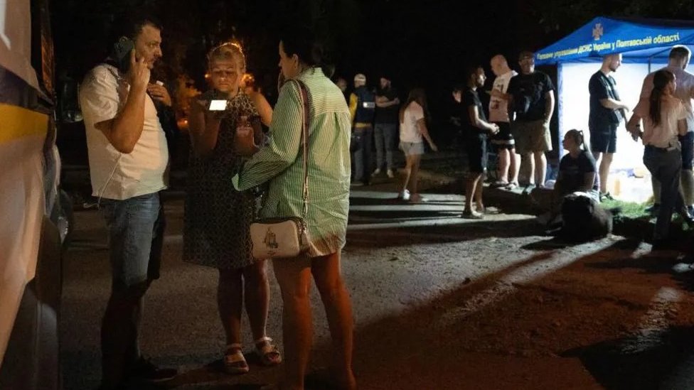 Ljudi napolju stoje i sede i telefoniraju tokom noći nakon napada Rusije na grad Poltavu u Ukrajini