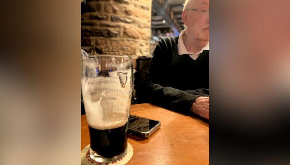 Četvrtak je za drugare i pivo: Nisu propustili okupljanje 56 godina 2 A close-up of a pint of Guinness that is only one third full, in front of a mobile phone. To the right in the background an elderly gentleman sits.