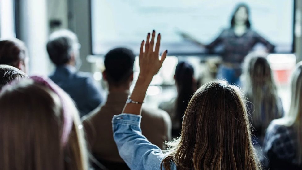 Zašto žene ređe postavljaju pitanja iz publike? 6 Žena drži predavanje ispred video ekrana dok druga žena u publici drži podignutu ruku da bi postavila pitanje