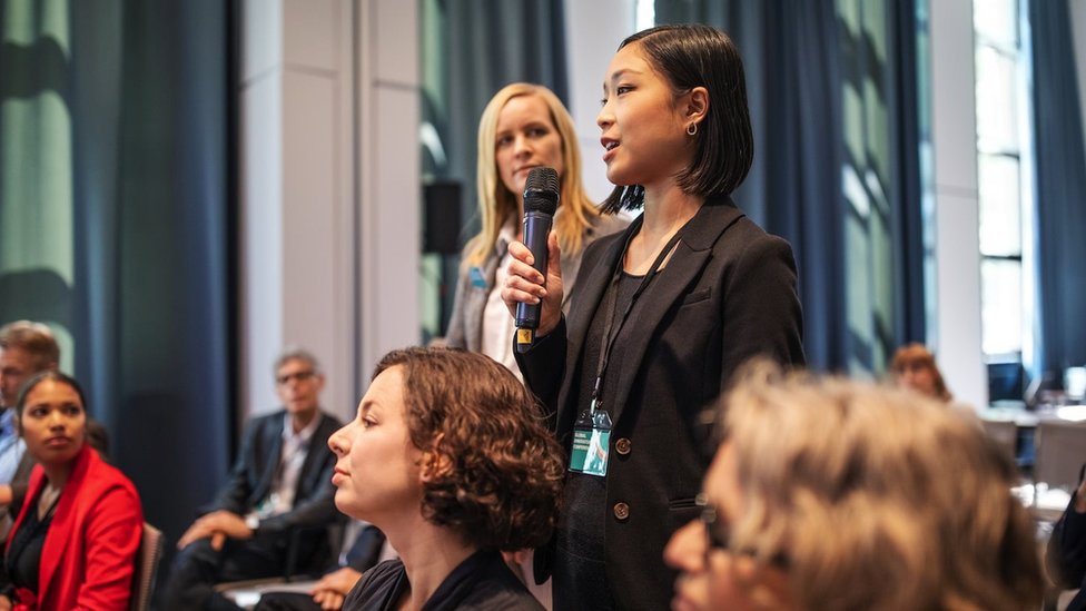 Zašto žene ređe postavljaju pitanja iz publike? 5 Žena postavlja pitanje stojeći i držeći mikrofon dok drugi ljudi sede i dok je druga žena gleda