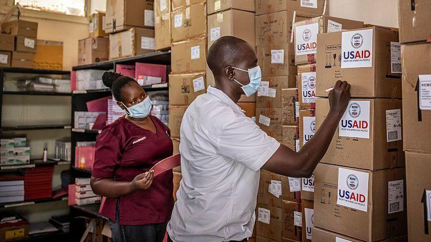 Trampovih 100 dana: Ko je najviše izgubio ukidanjem američke pomoći 5 Muškarac i žena sa maskama za lice u prostoriji sa mnoštvo kartonskih kutija nagomilanih jedna na drugu, na kojima piše USAID