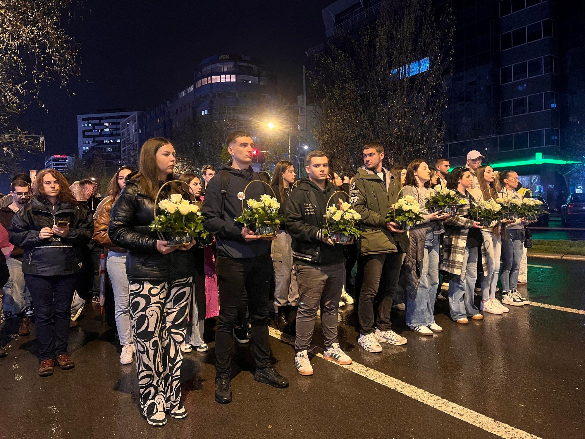 Mladi ljudi drže bukete belih ruža na kiši tokom noći, u koloni