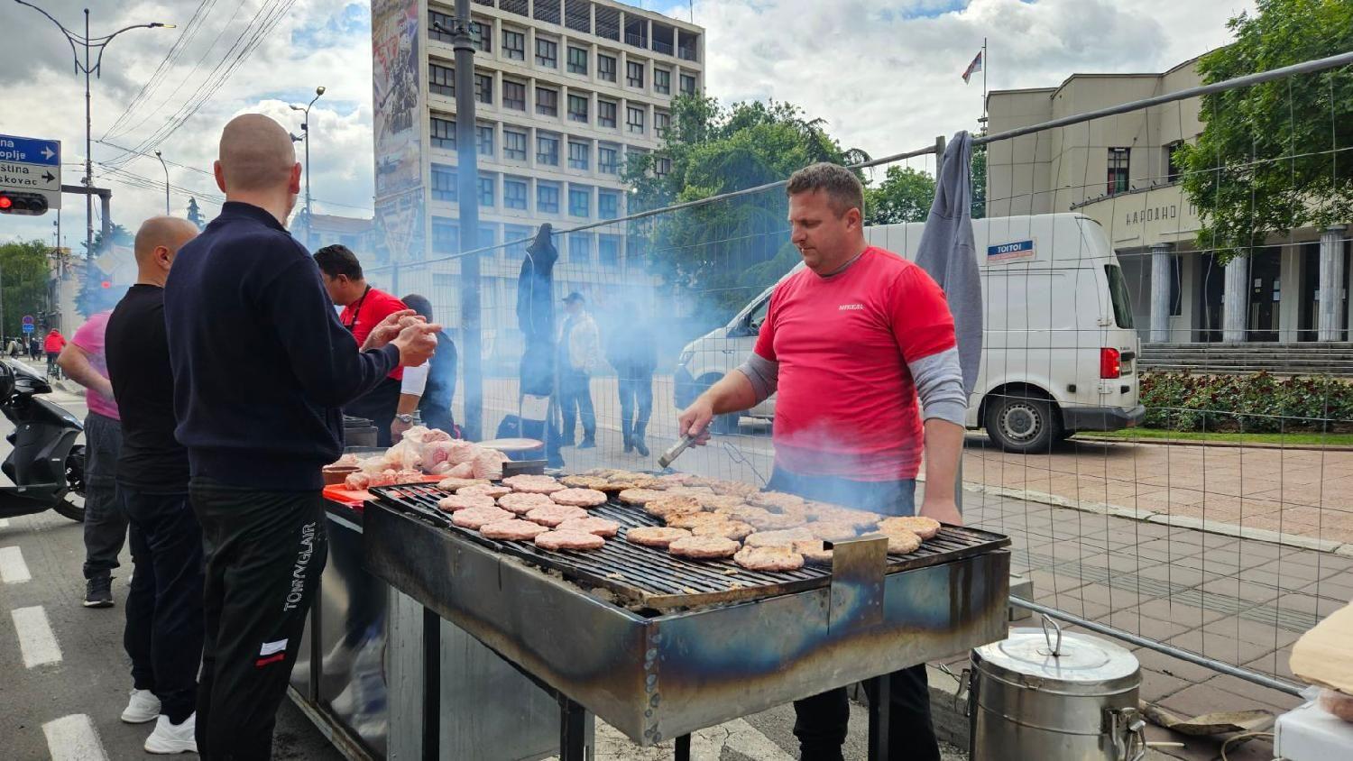 Muškarac u crvenoj majici okreće pljeskavice na roštilju na trgu