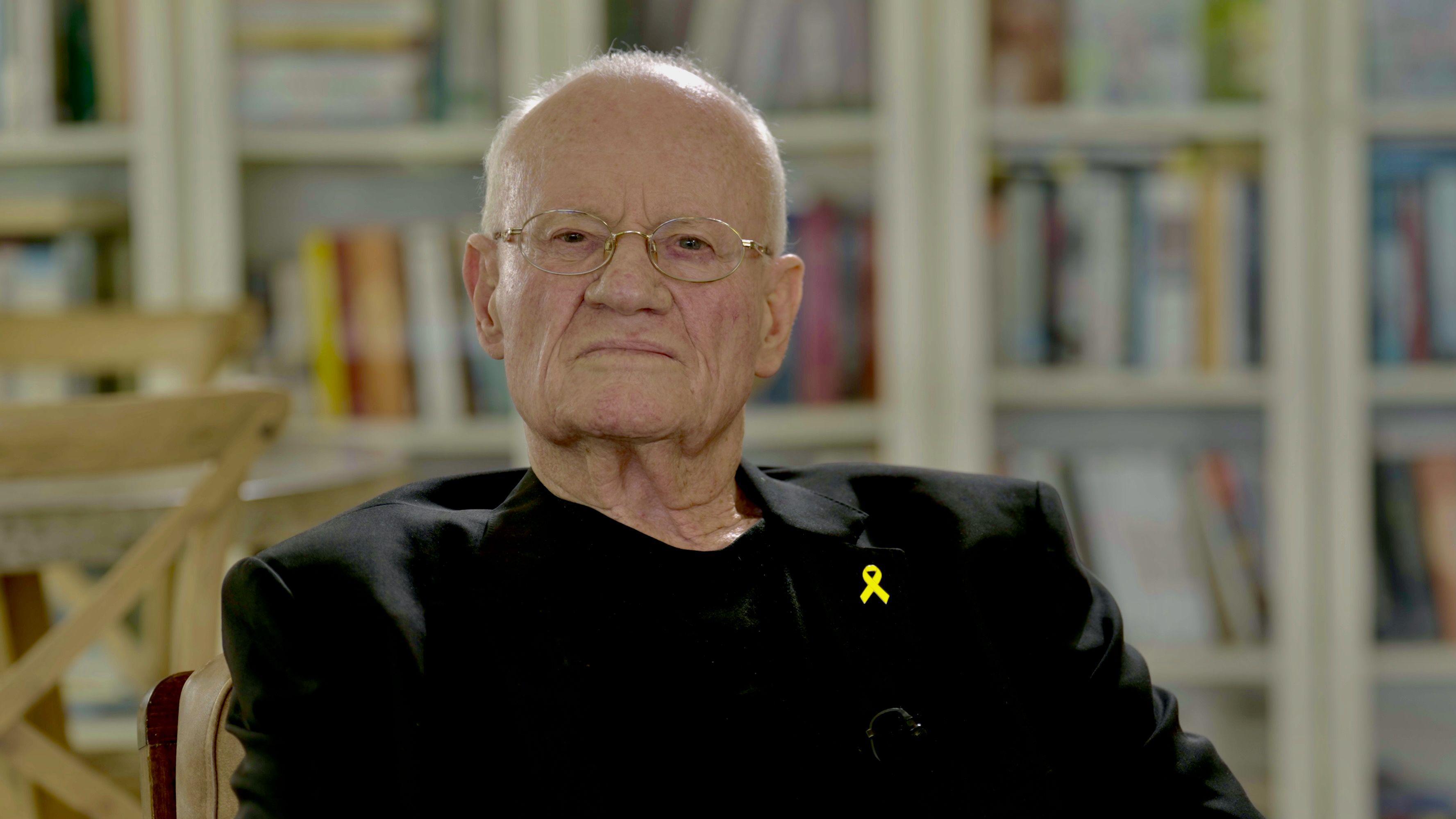 Danny Yatom sits in an office, in a black shirt and jacket with glasses on, with books in the background