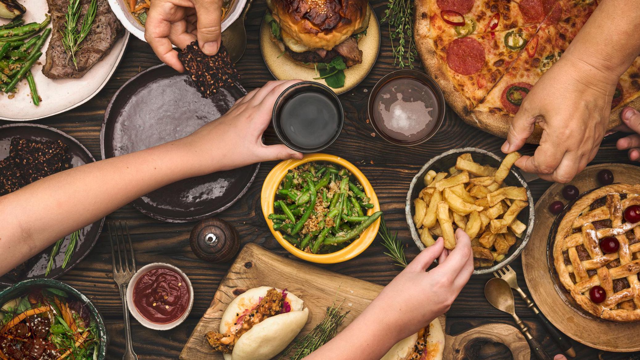 Čeka li nas decenija rekordnih cena hrane 1 Ruke različitih ljudi biraju hranu, pomfrit, picu, salate i meso, iz činija na drvenom stolu