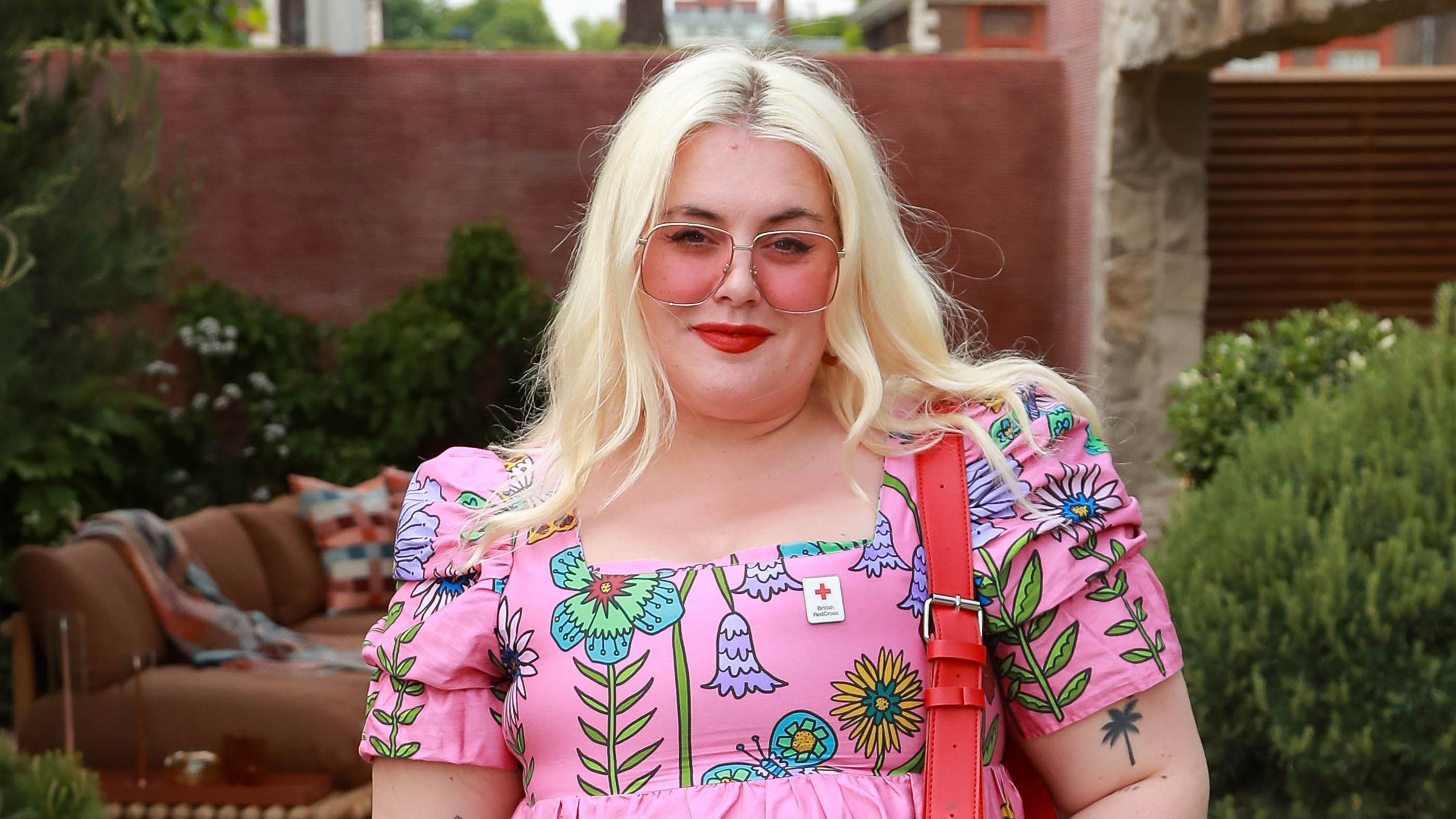 Plavuša sa sunčanim pink naočalama i u roze haljini na cvetove gleda u kameru