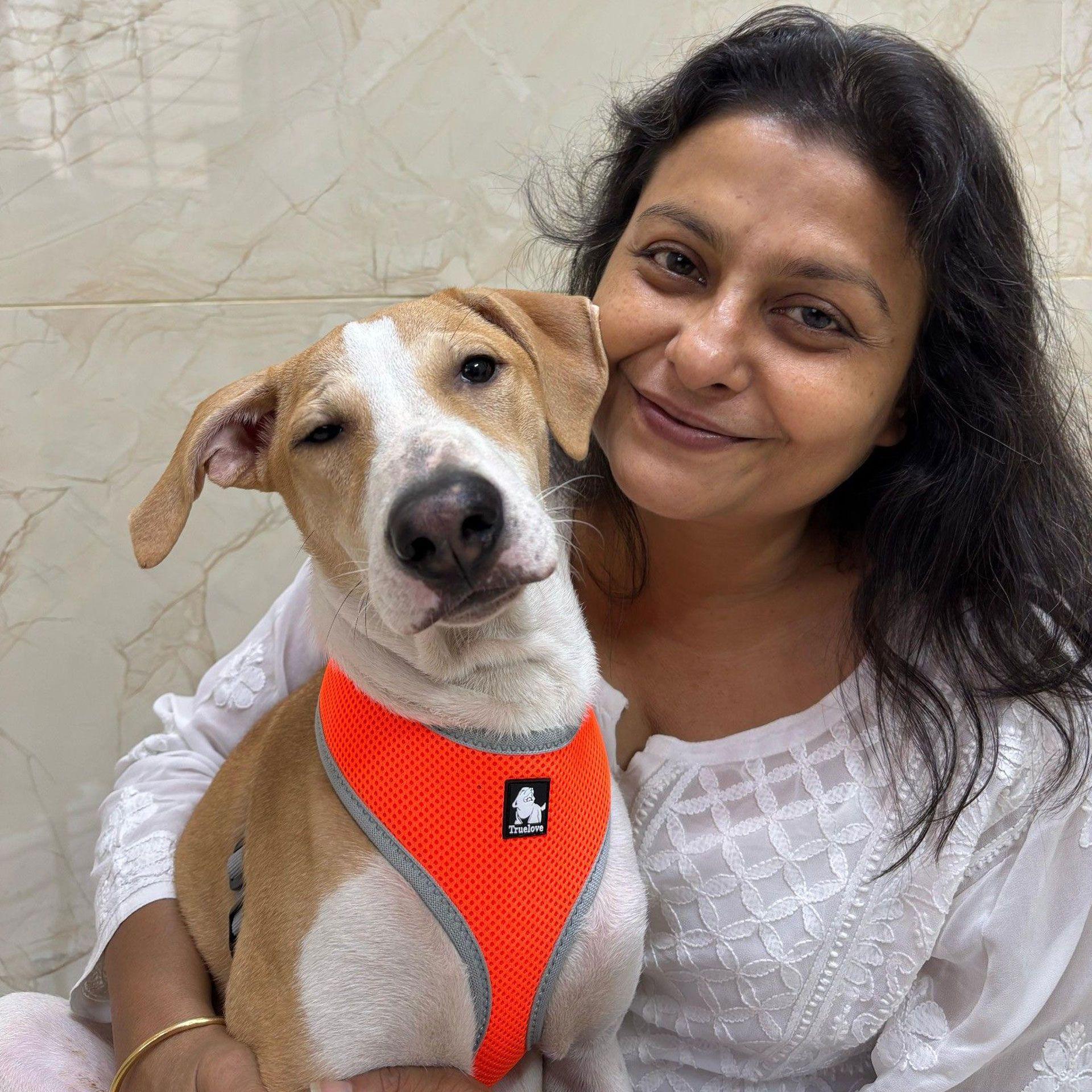 Žena sa dugom tamnom kosom sedi i grli smeđe-belog psa.
