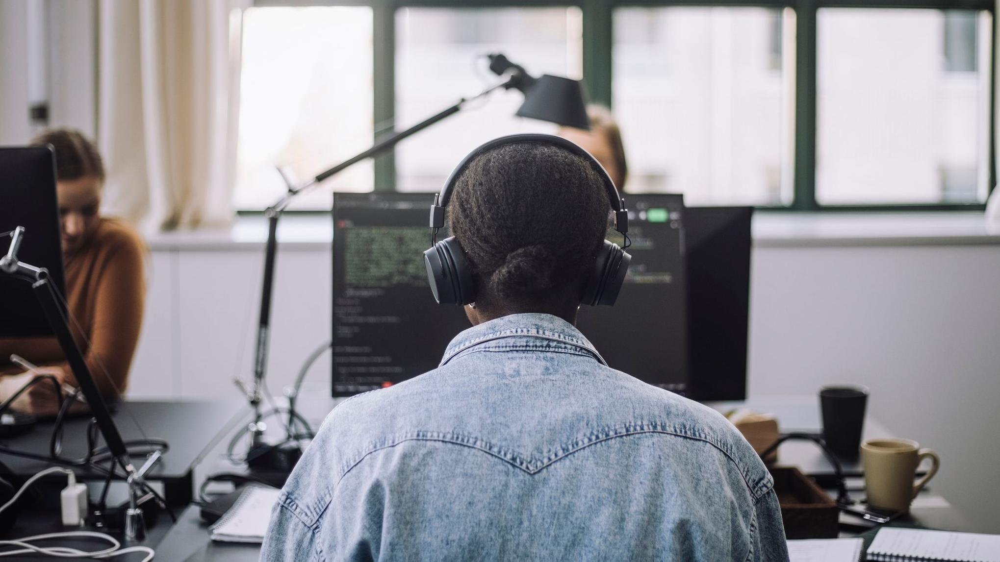 Žena u teksas jakni slikana s leđa dok radi za računarom i ima slušalice na ušima 