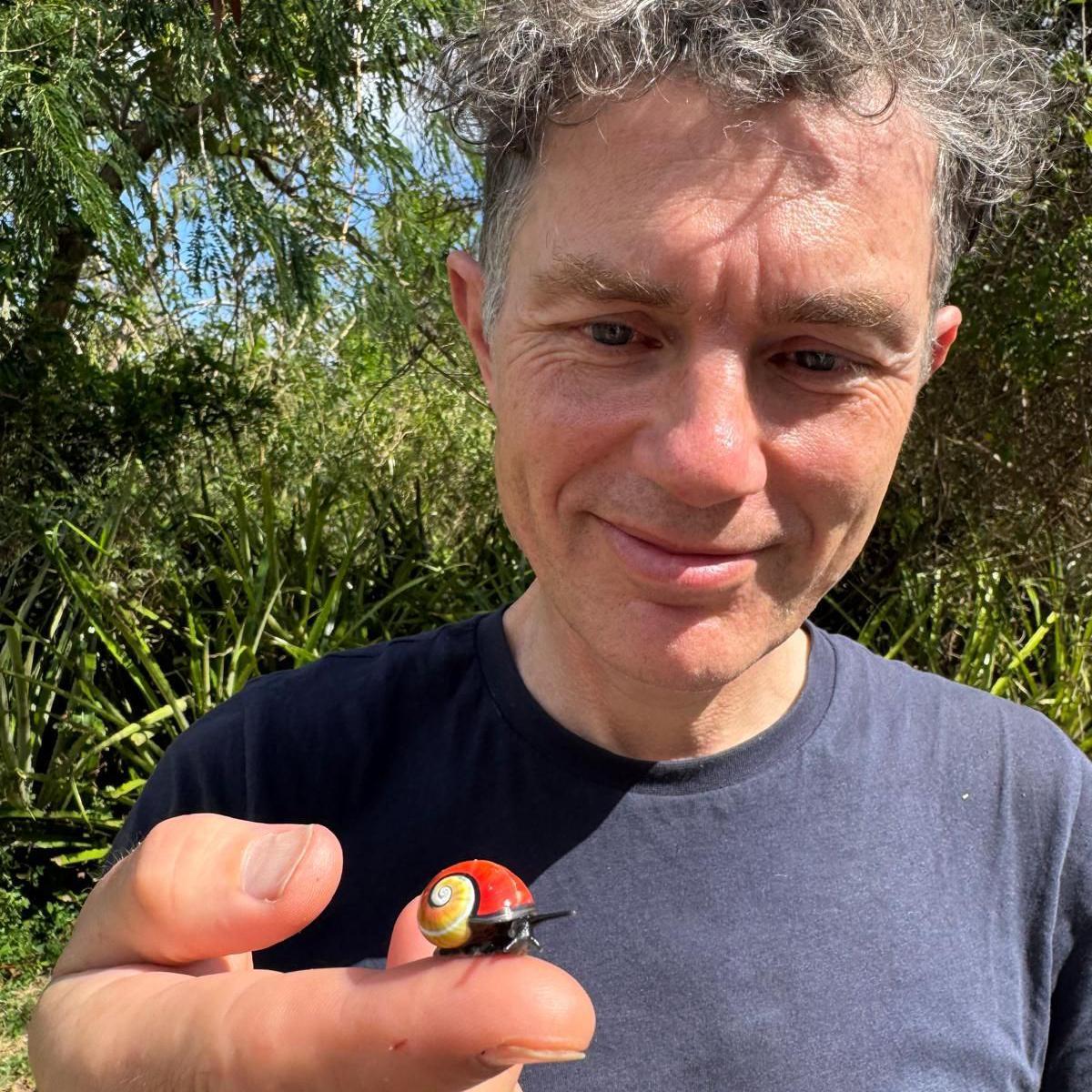 A smiling man in a navy blue T-shirt holds a brightly coloured snail towards the camera