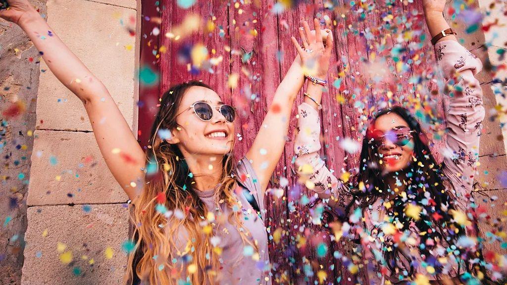 Dve devojke duge plave i tamne kose sa rukama u vazduhu, smeju se, na njih padaju šarene konfete, obe imaju naočale za sunce