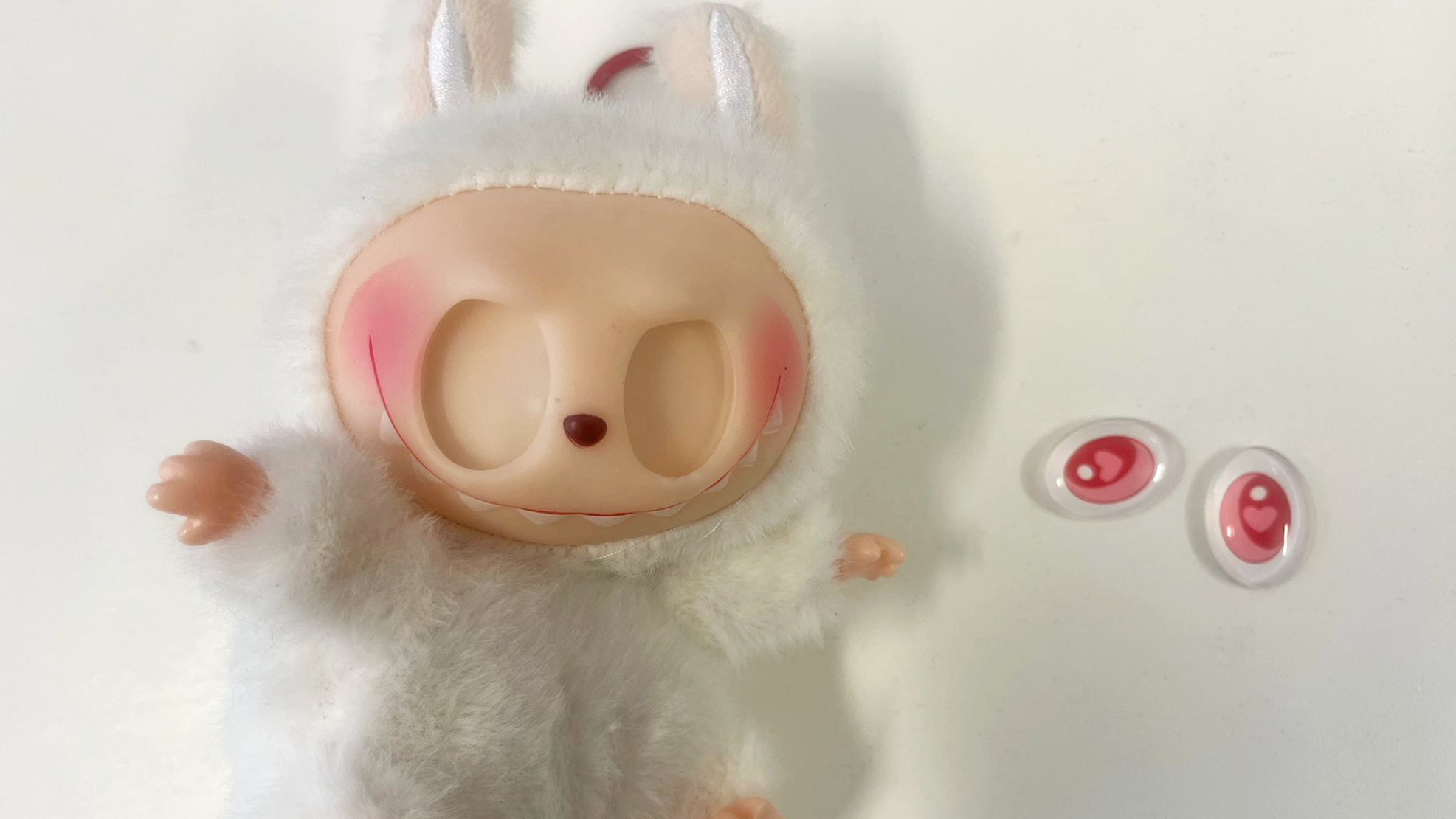 Bela pahuljasta lutka Labubu sa praznim očnim dupljama. Njene velike ružičaste oči sede pored nje na stolu.
