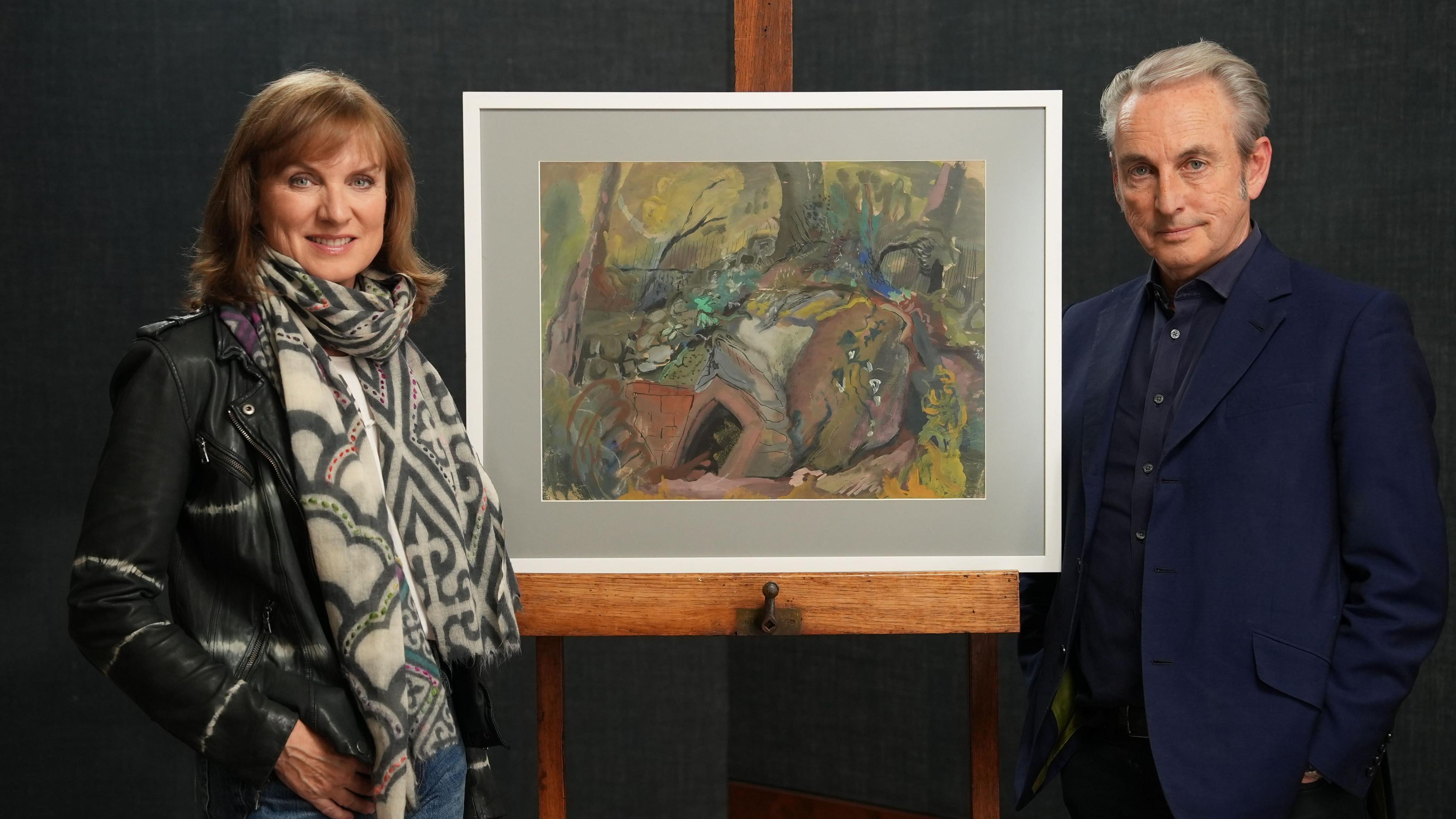 Muškarac i žena stoje ispred uramljene slike na štafelaju. Ona nosi crnu kožnu jaknu i šareni šal. On nosi tamnoplavu košulju i plavi sako.