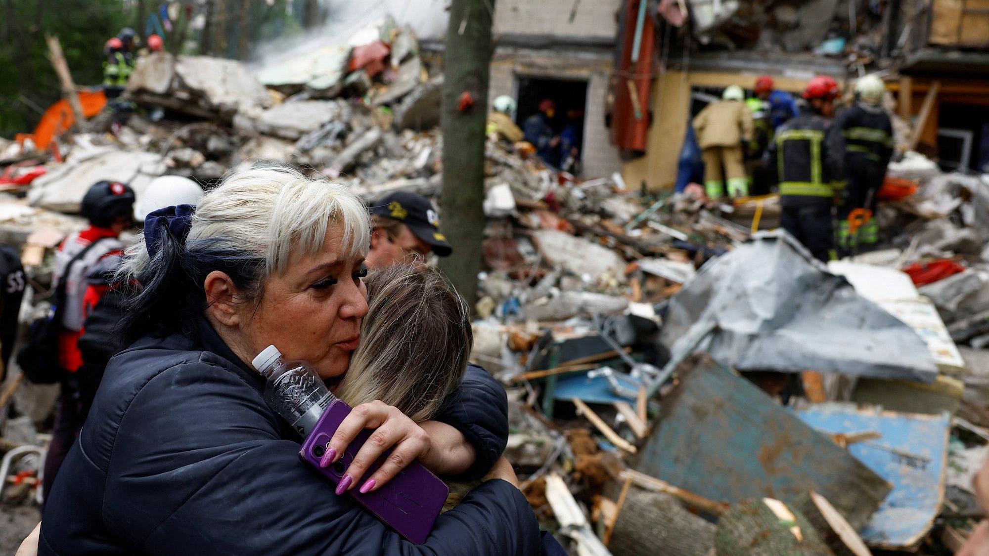 Dan žalosti u Kijevu posle desetina mrtvih i ranjenih u ruskim napadima 4 Žena u jakni, sede kose, grli devojčicu svetlije kose i drži u ruci telefon sa ljubičastom maskom i flašicu vode, oko njih su ruševine, a u daljini se vide spasilačke službe