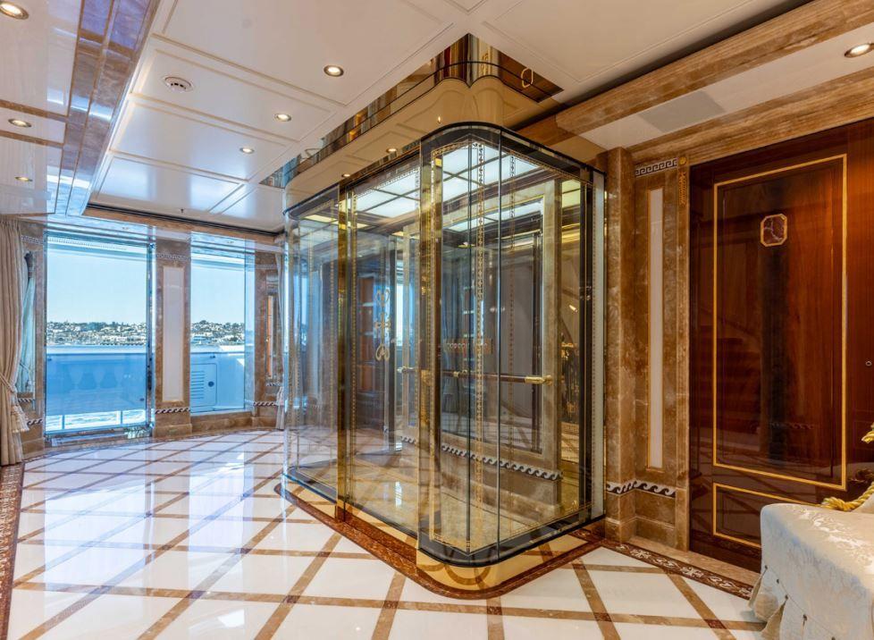 'Beskonačni' bazen, helidrom, osam soba: 'Superjahta ruskog milijardera' na aukciji 4 Lift sa zlatnim okvirom i staklenim zidovima, smešten uz zid. Pod je beo i sjajan sa pravougaonim smeđim ivicama. More i obala se vide kroz prozore u pozadini.